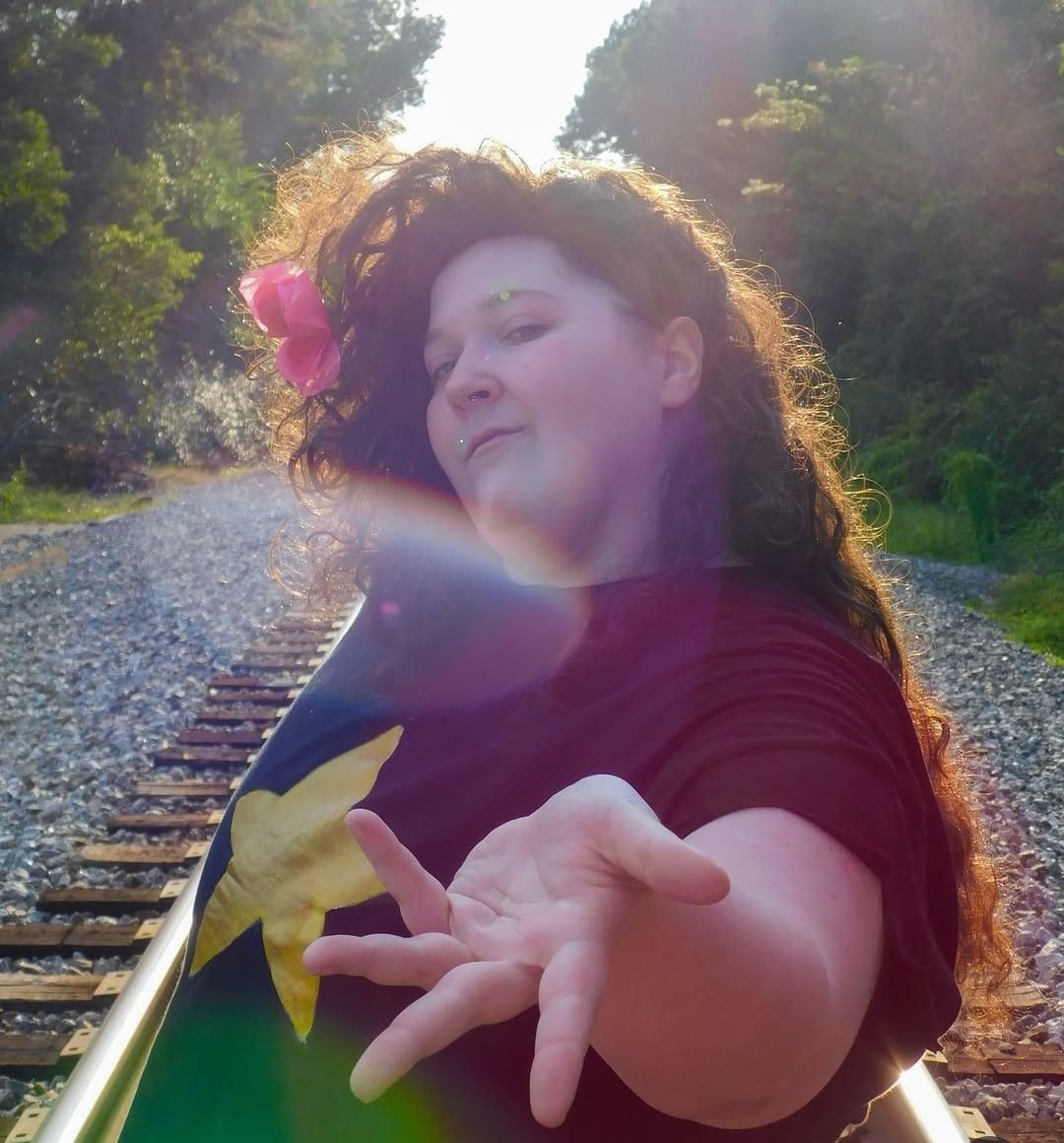 An image of a plus-size nonbinary person dressed as Greg Universe from Steven Universe. They are standing at a 3/4 angle and are gesturing their hand toward the camera.