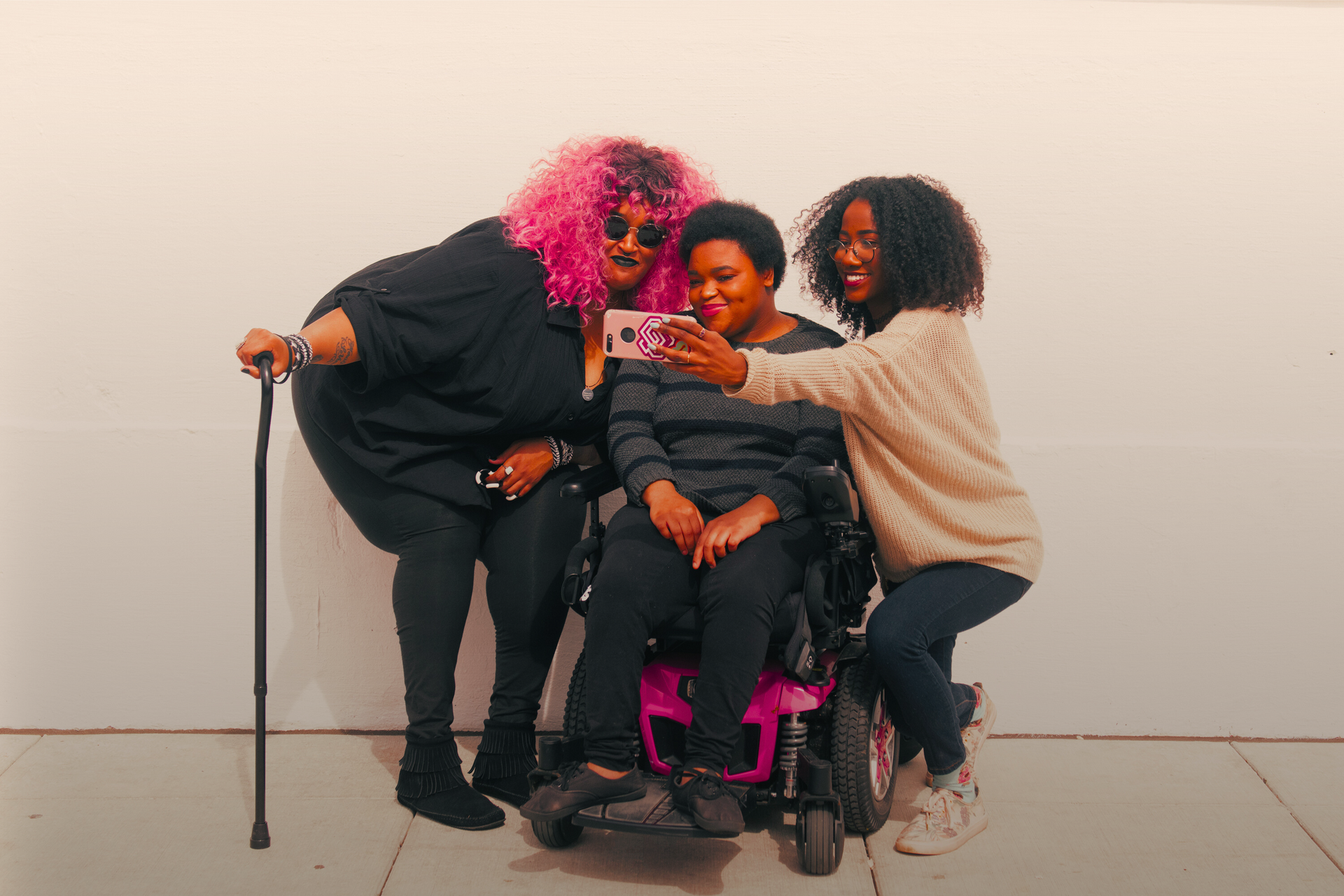 A zoomed out full body shot showing three Black and disabled friends (a non-binary person with a cane and tangle stim toy, a non-binary person sitting in a power wheelchair, and an invisibly disabled femme) smiling and taking a cell phone selfie together. All are outdoors in front of a white wall.

Image Credit: https://affecttheverb.com/collection/