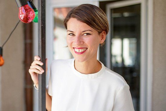 Woman smiling, wearing white shirt, standing in doorway with string lights.