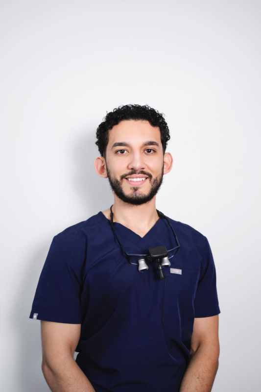 Man in dark blue scrubs smiles at the camera, dental loupes around his neck, against a white background.