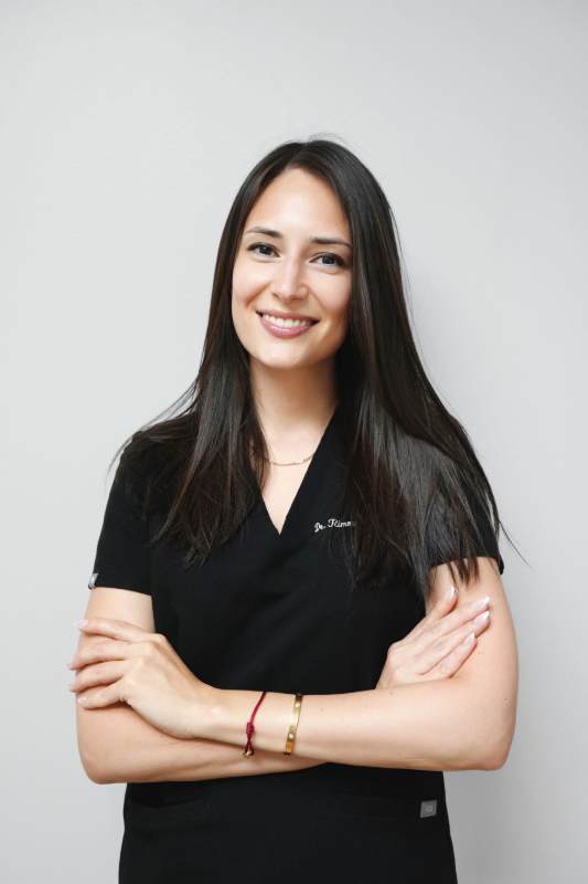 Woman with dark hair, wearing black scrubs, arms crossed, smiling. Plain, light background.