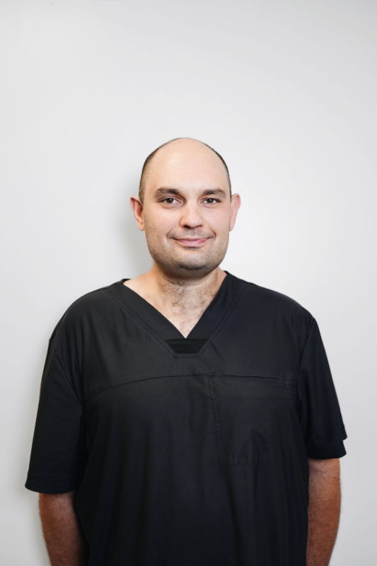 Man in black scrubs smiles at the camera, against a white wall.