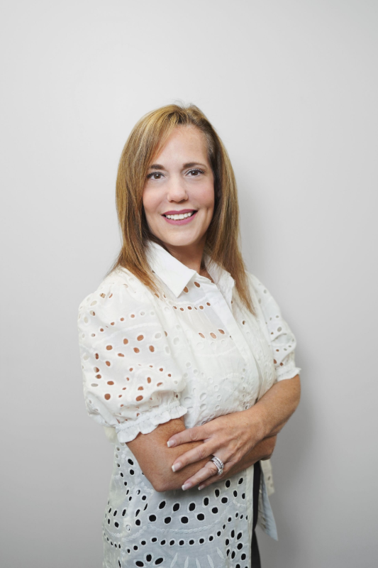 Woman with blond hair smiles, arms crossed, wearing a white eyelet shirt.