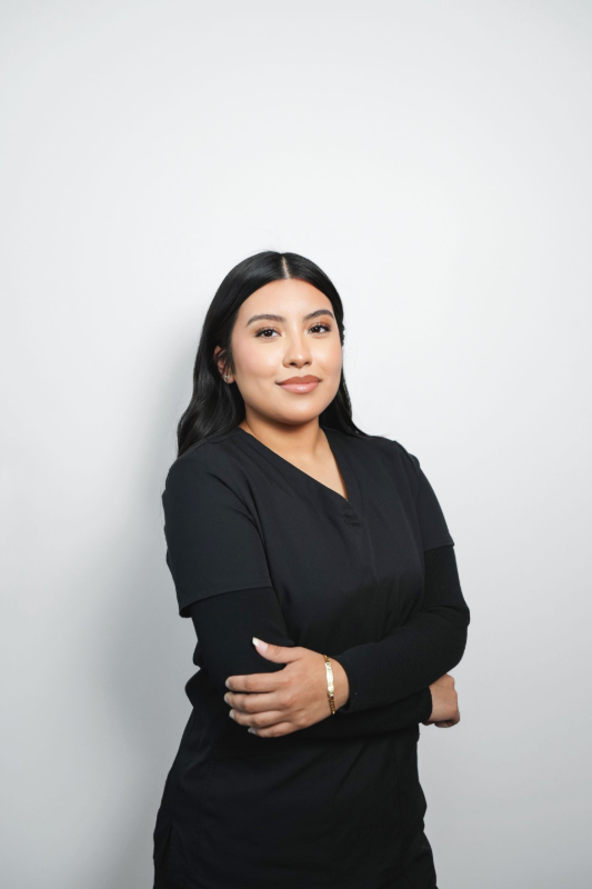 Woman in black scrubs, arms crossed, smiles at the camera against a white backdrop.
