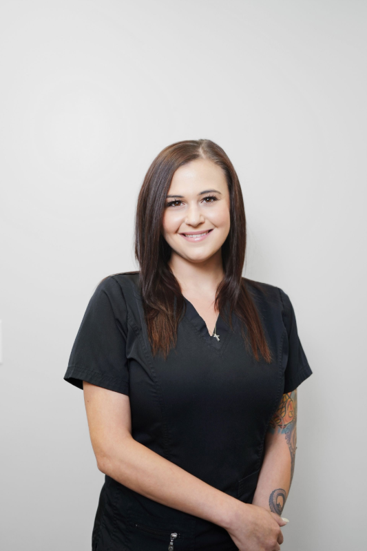 Woman in black scrubs smiling, standing against a light gray wall.