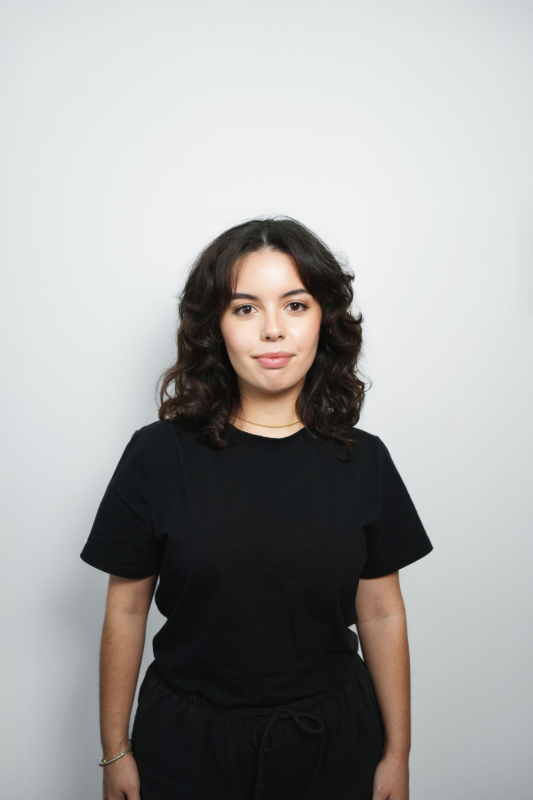 Woman with dark curly hair, wearing a black t-shirt, standing against a white backdrop.