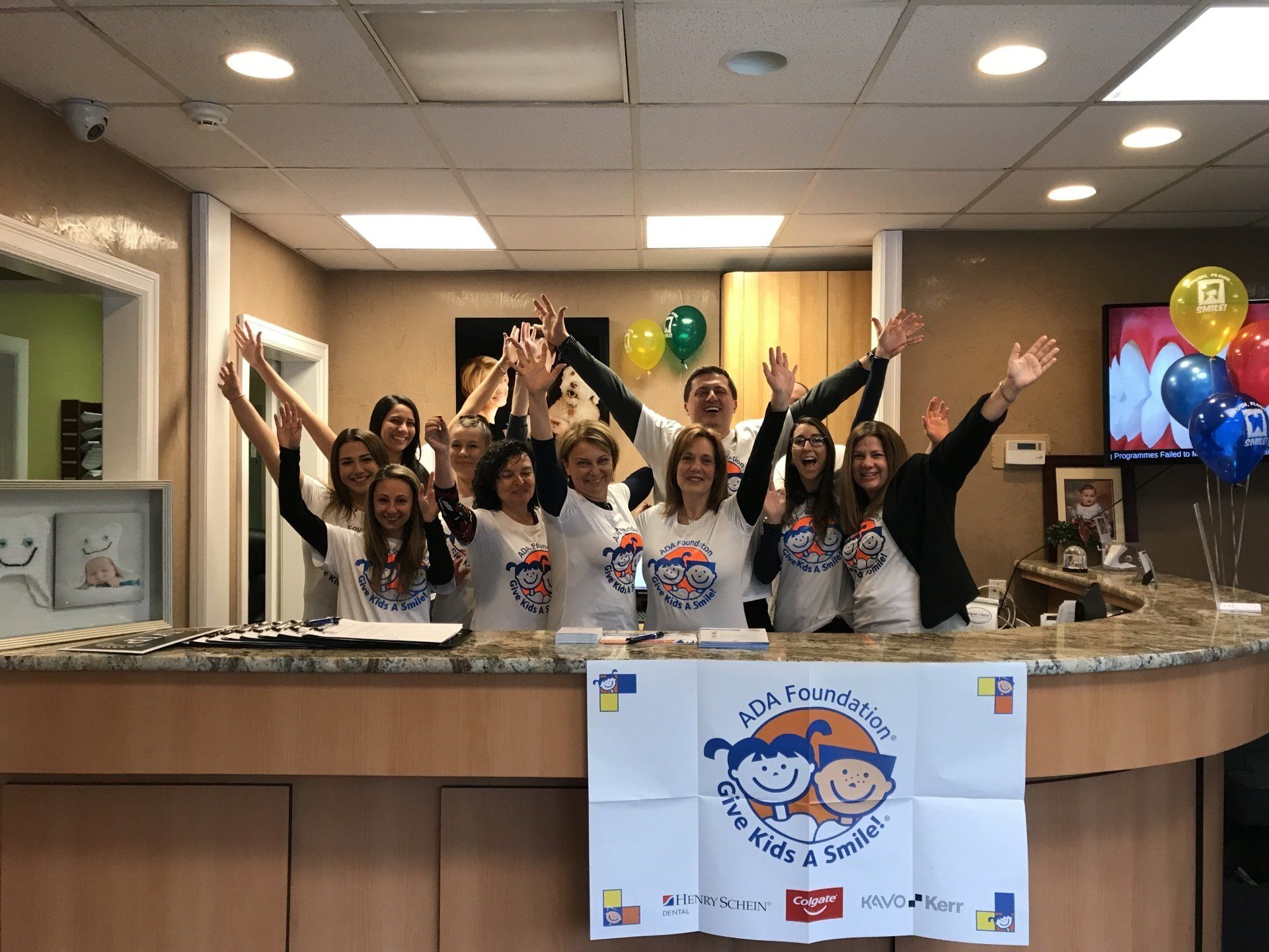 Dental office staff with raised arms, smiling. ADA foundation sign. Balloons decorate the desk.