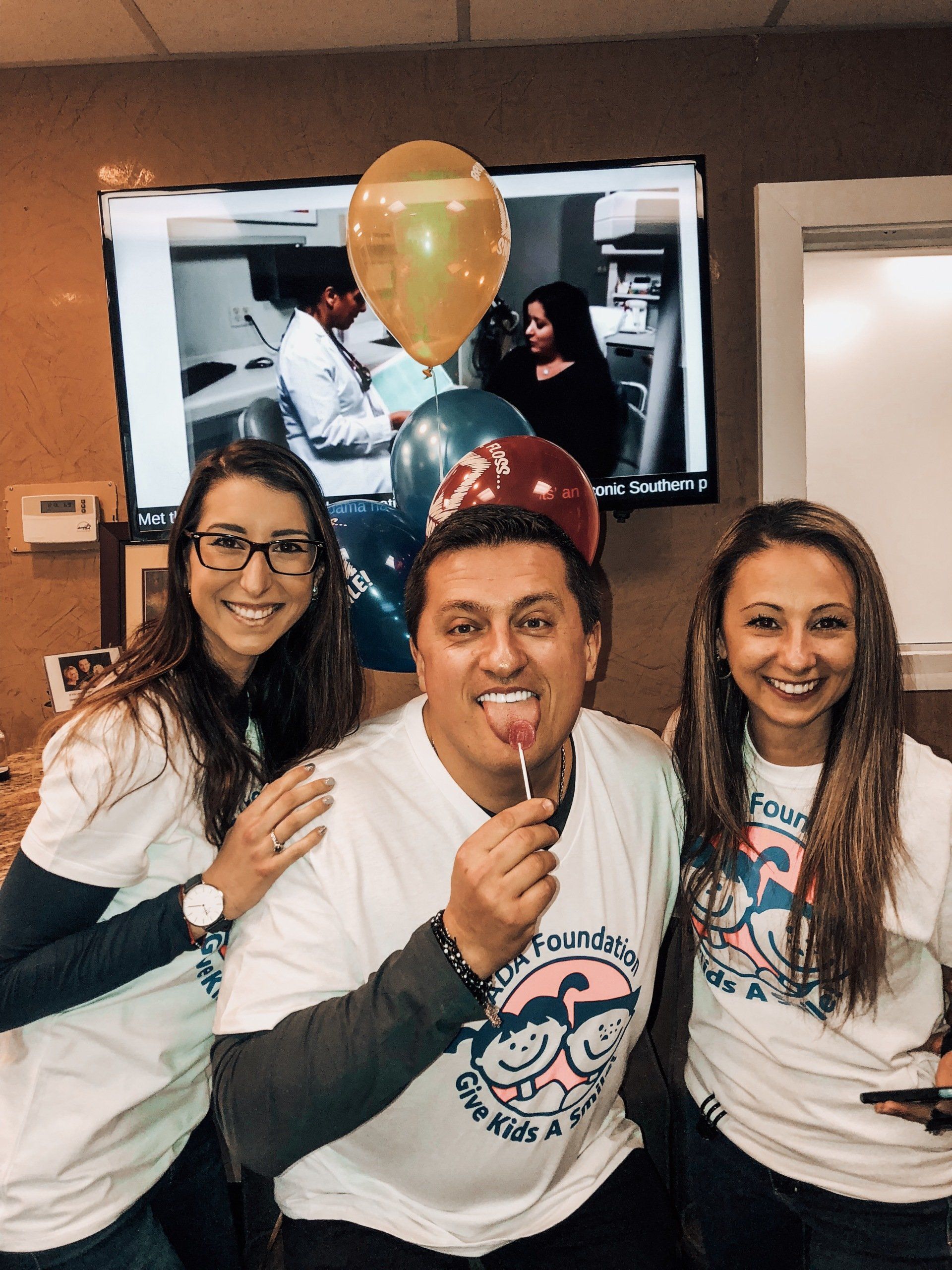 Three people smiling with balloons; one licking a lollipop. White t-shirts in a dental office.