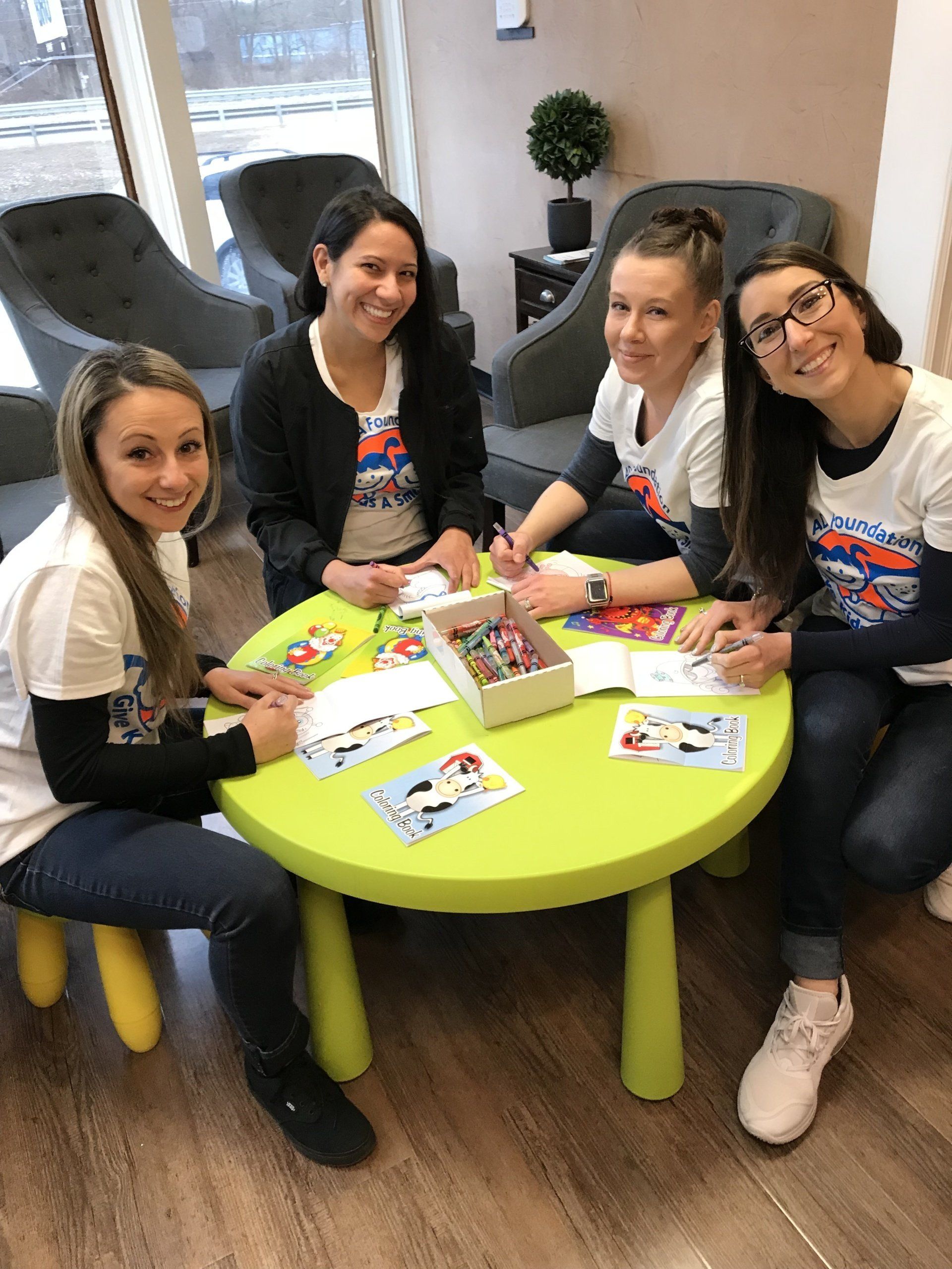 Four smiling women at a green table, coloring. They wear white shirts and jeans, in a waiting room.