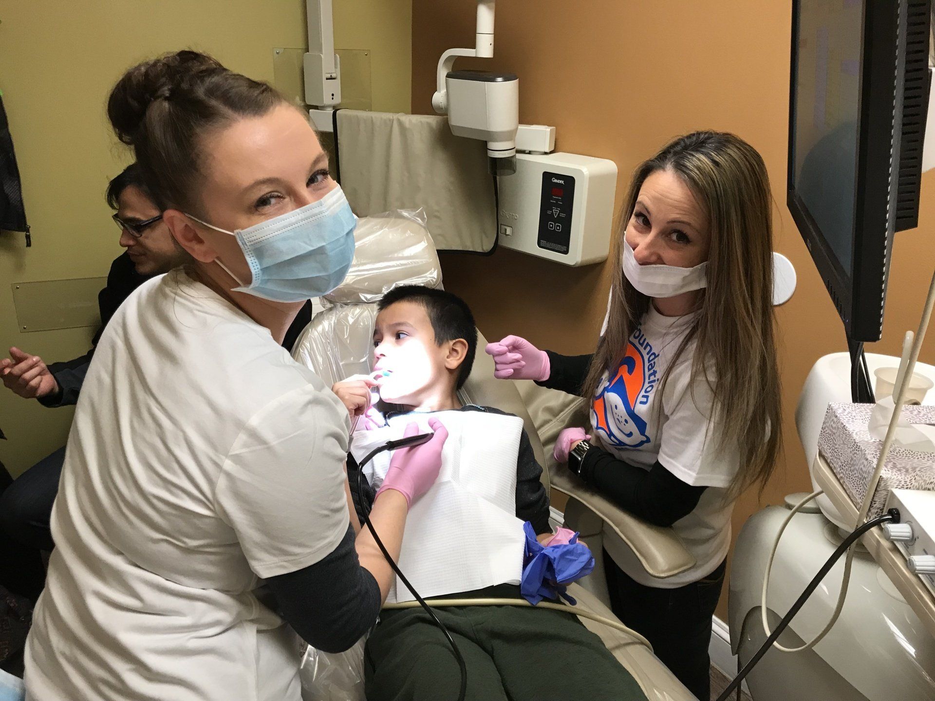 Two dental staff assist a child in a dentist's chair, both wearing masks.