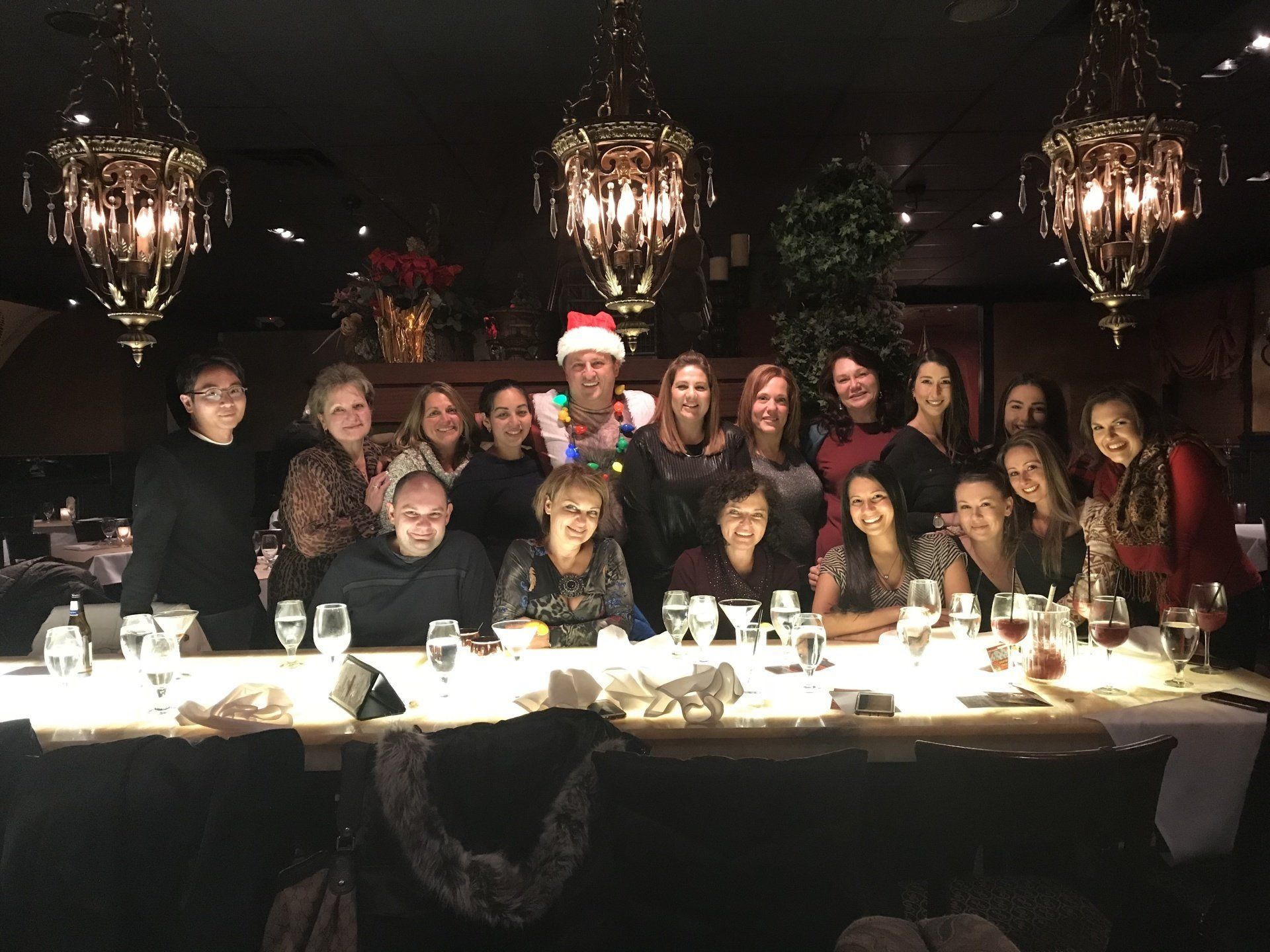 A group of people smiling at a long table in a restaurant, festive lights and decorations.