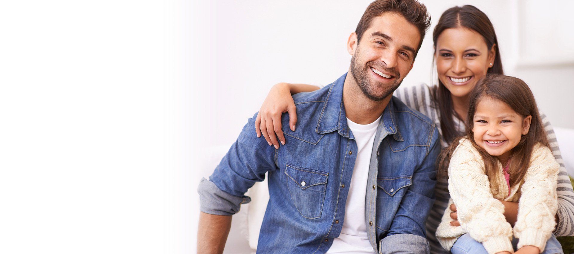 Family of three smiling, father with arm around mother, daughter in front. White background.