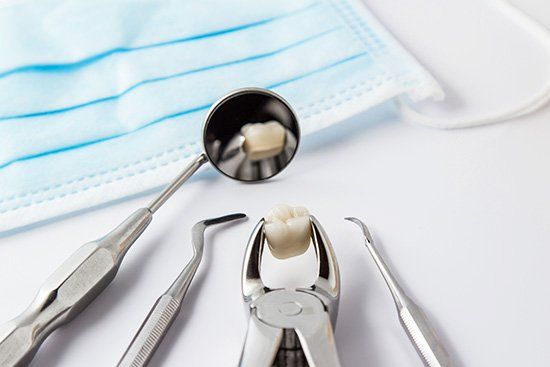 Dental tools arranged on a white surface: mirror, pick, pliers holding a tooth, and a face mask.