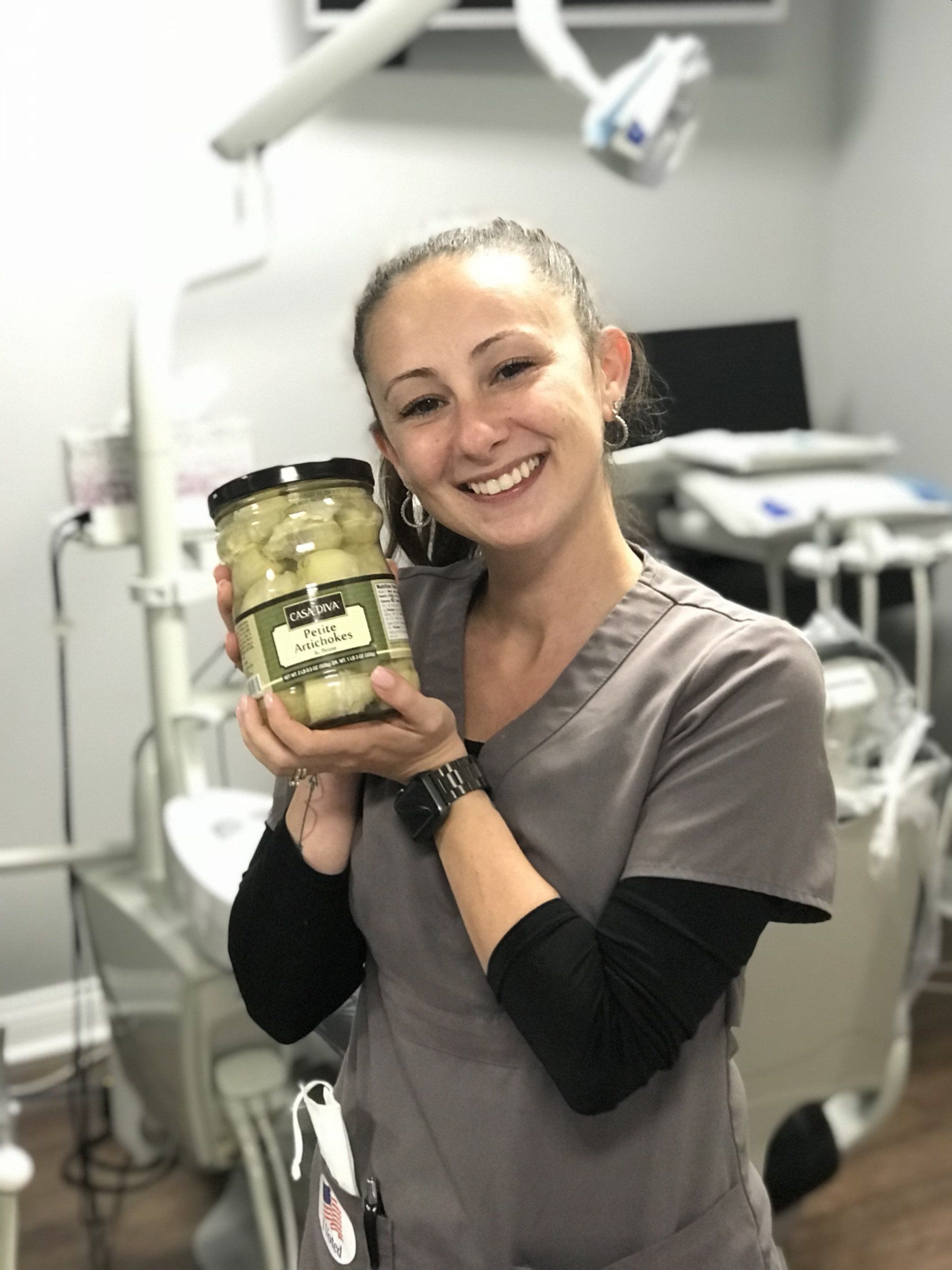 Woman in scrubs smiles, holding a jar of artichoke hearts in a dental office.
