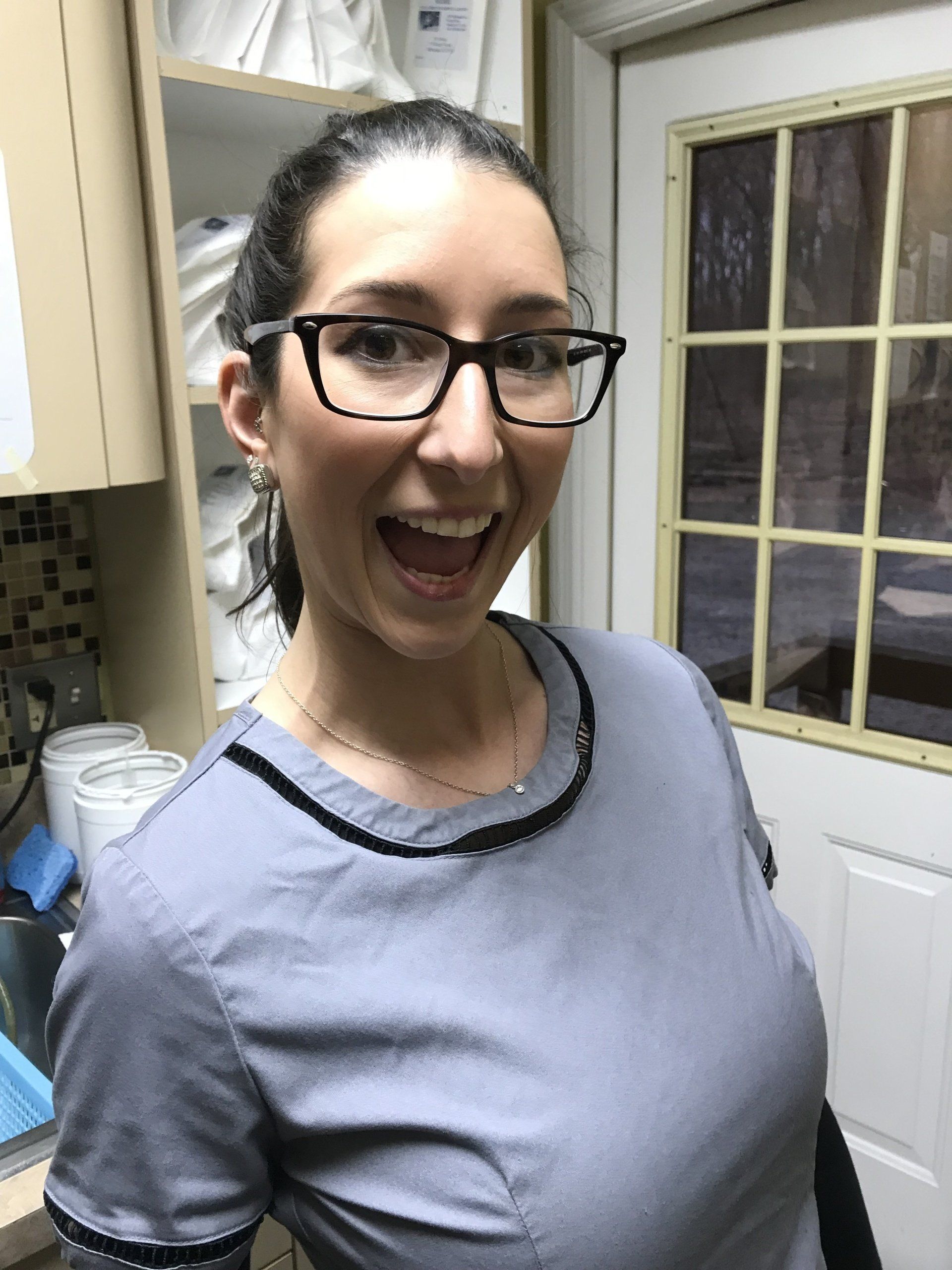 Woman wearing glasses and a gray scrub top smiles broadly in a medical office setting.