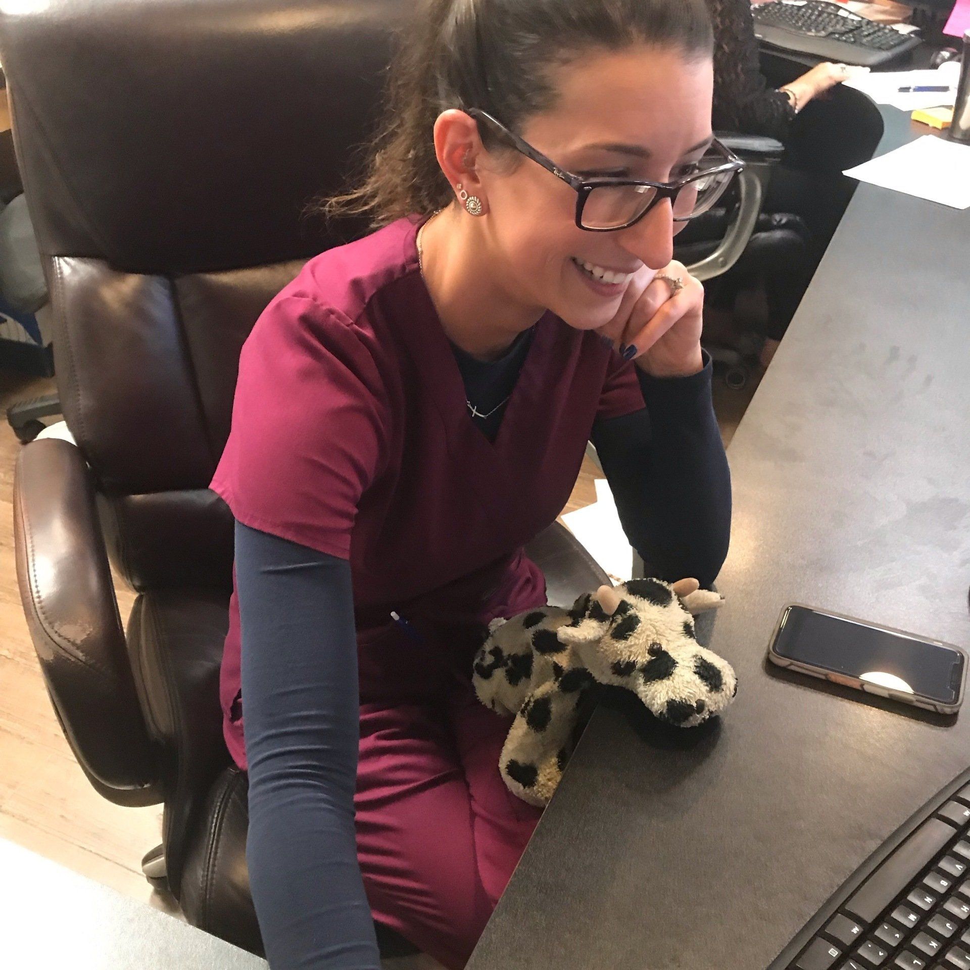 Woman in burgundy scrubs smiles at a desk. Cow stuffed animal, phone, and keyboard visible.