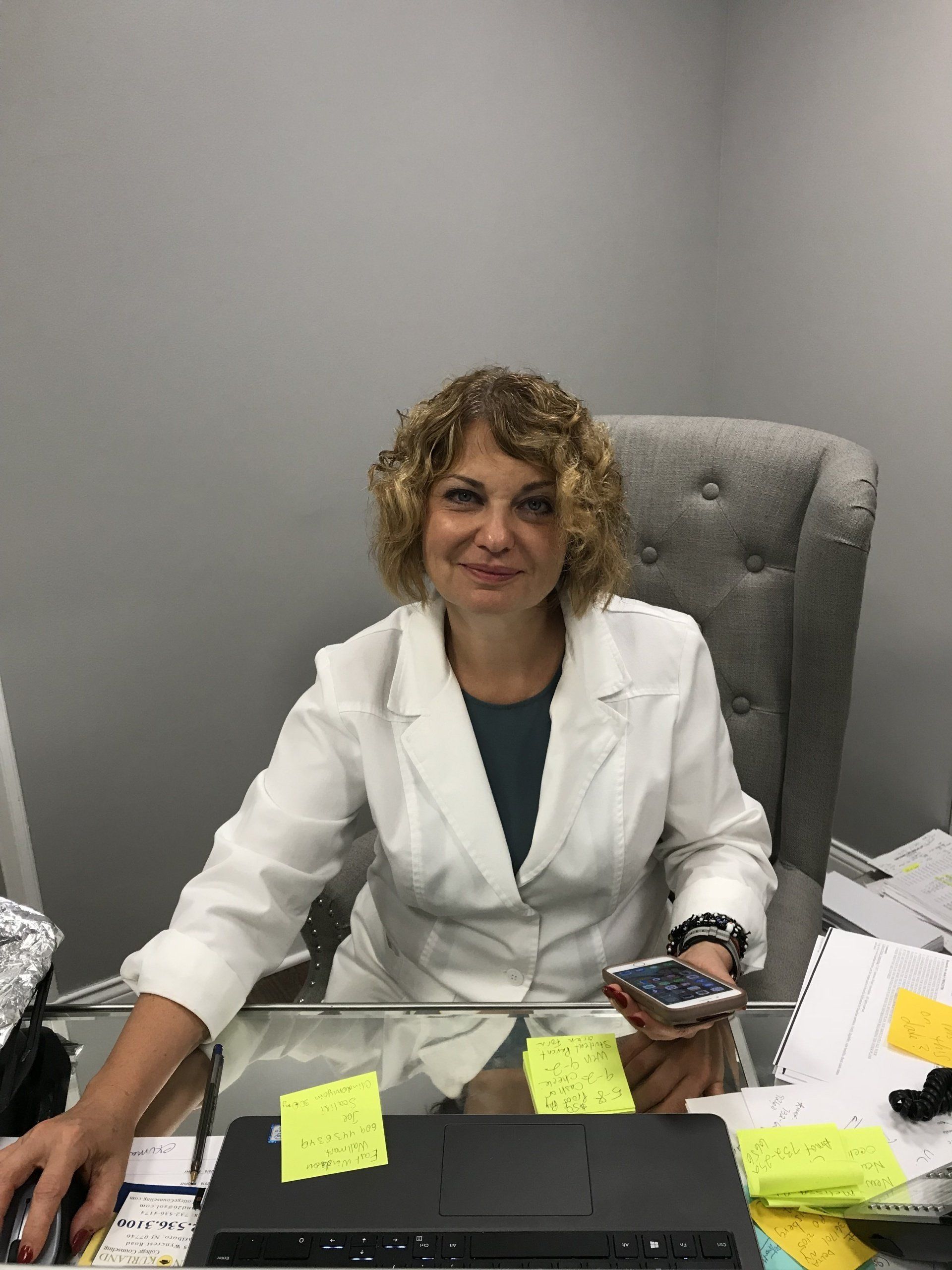 Woman with curly blonde hair sits at desk, wearing a white blazer, holding phone.