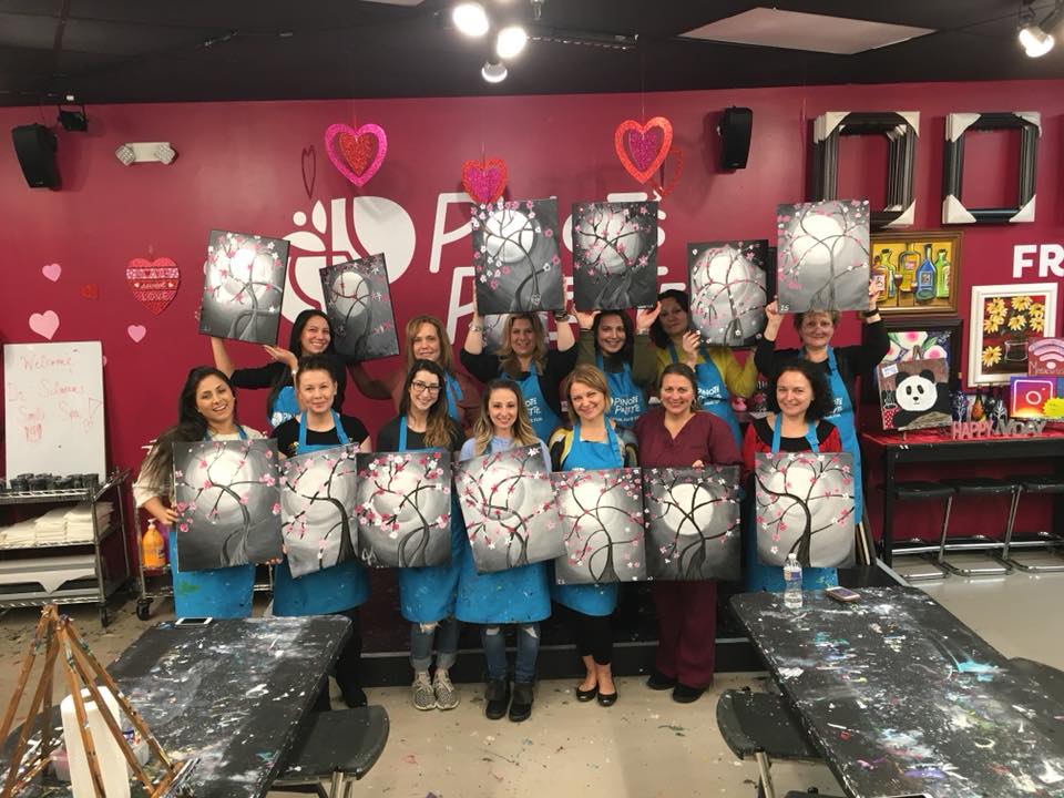 Group of women at a painting class holding up their completed art of a moonlit tree, in a studio setting.