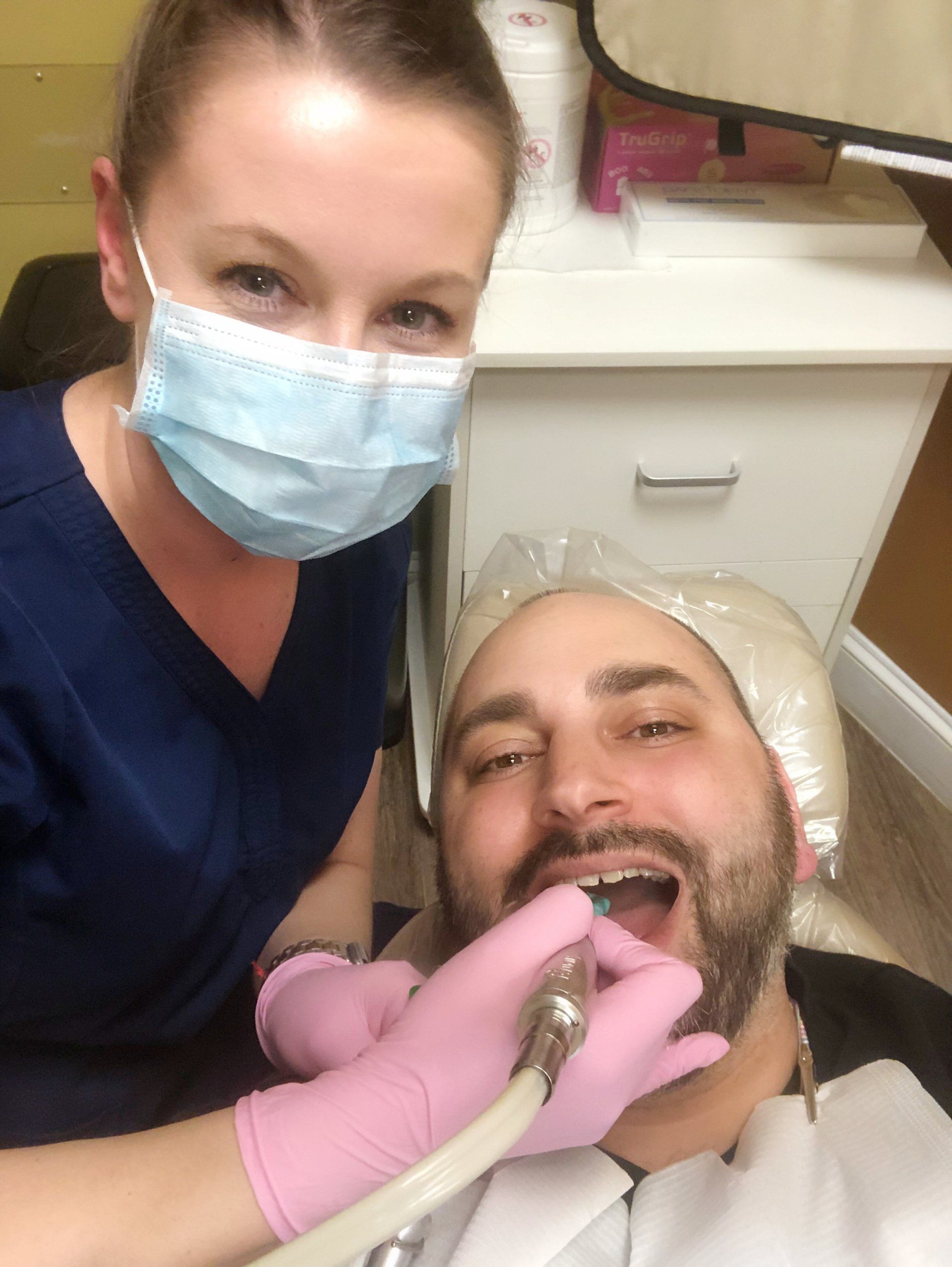 Dentist in blue scrubs and mask, with pink gloves, working on a man's teeth.