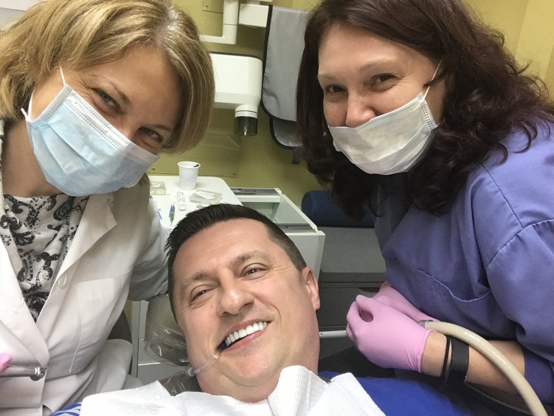 Man smiles in dental chair as two dental professionals smile, wearing masks and gloves.
