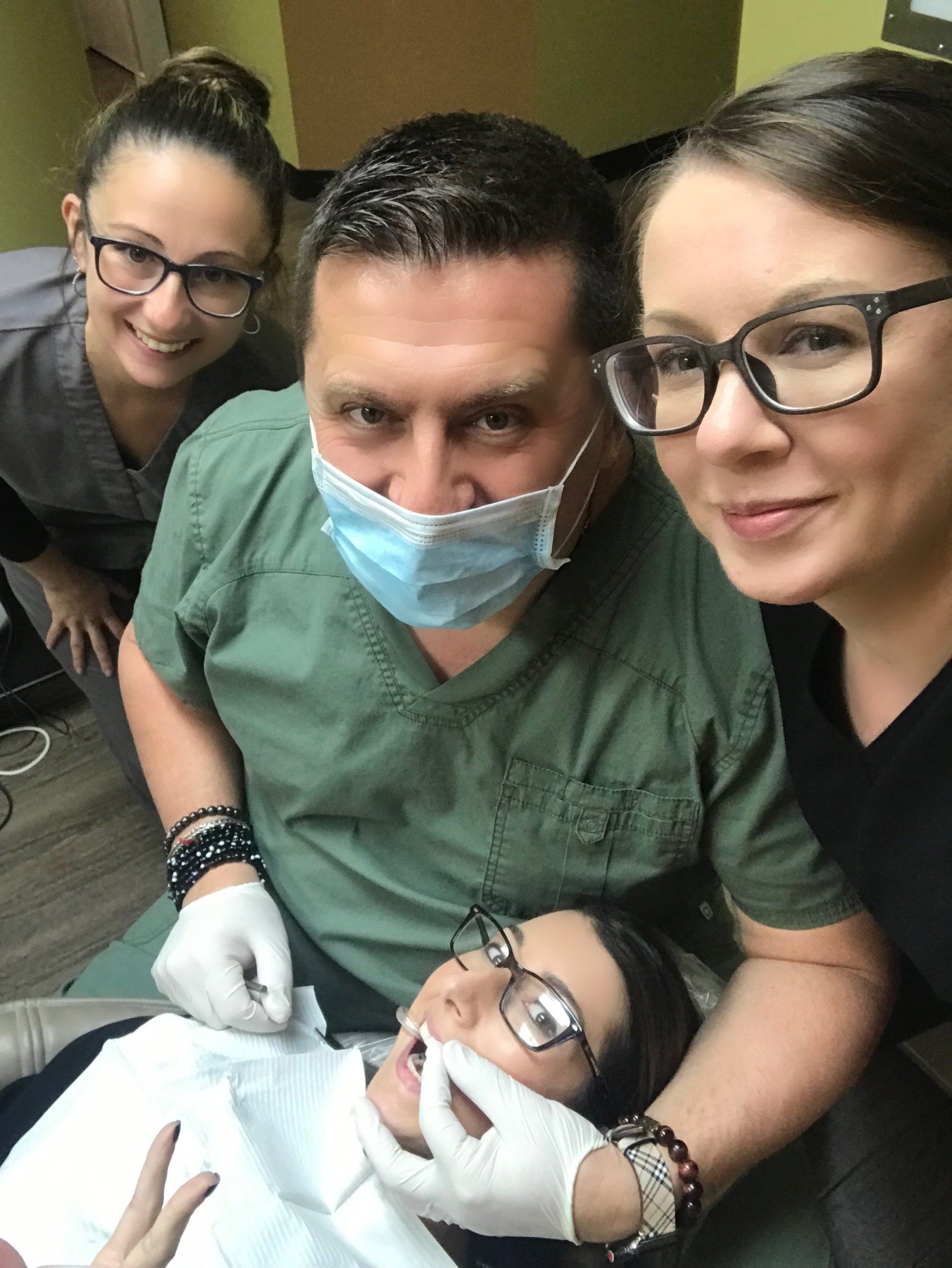 Dentist and assistants with patient in dental chair; all wearing masks, gloves, and glasses.