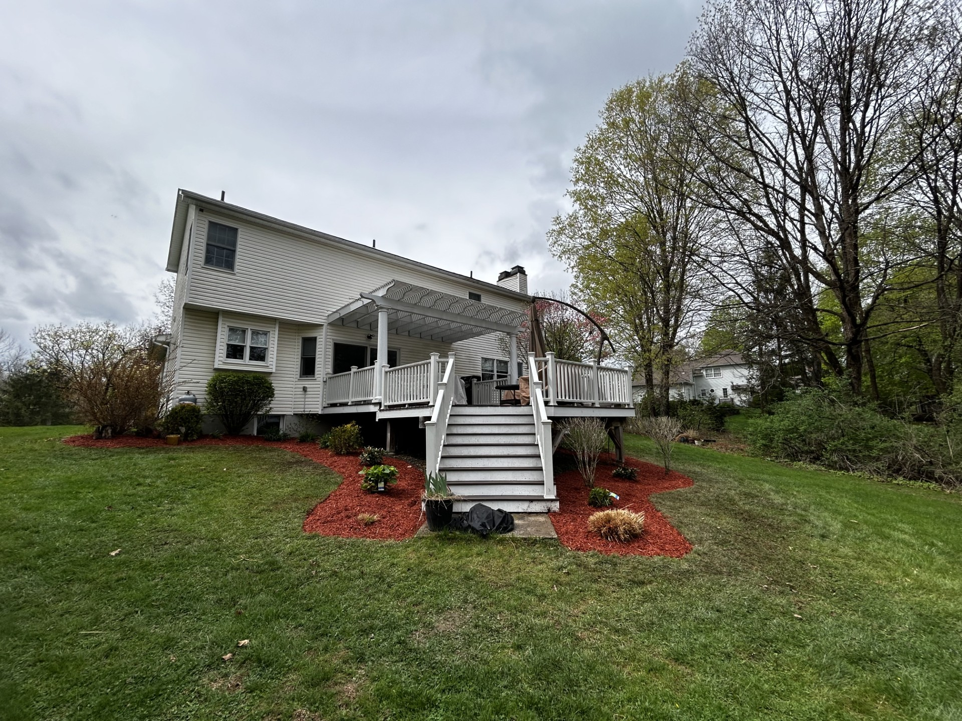 White house with a deck and steps, surrounded by green grass and trees under a cloudy sky.