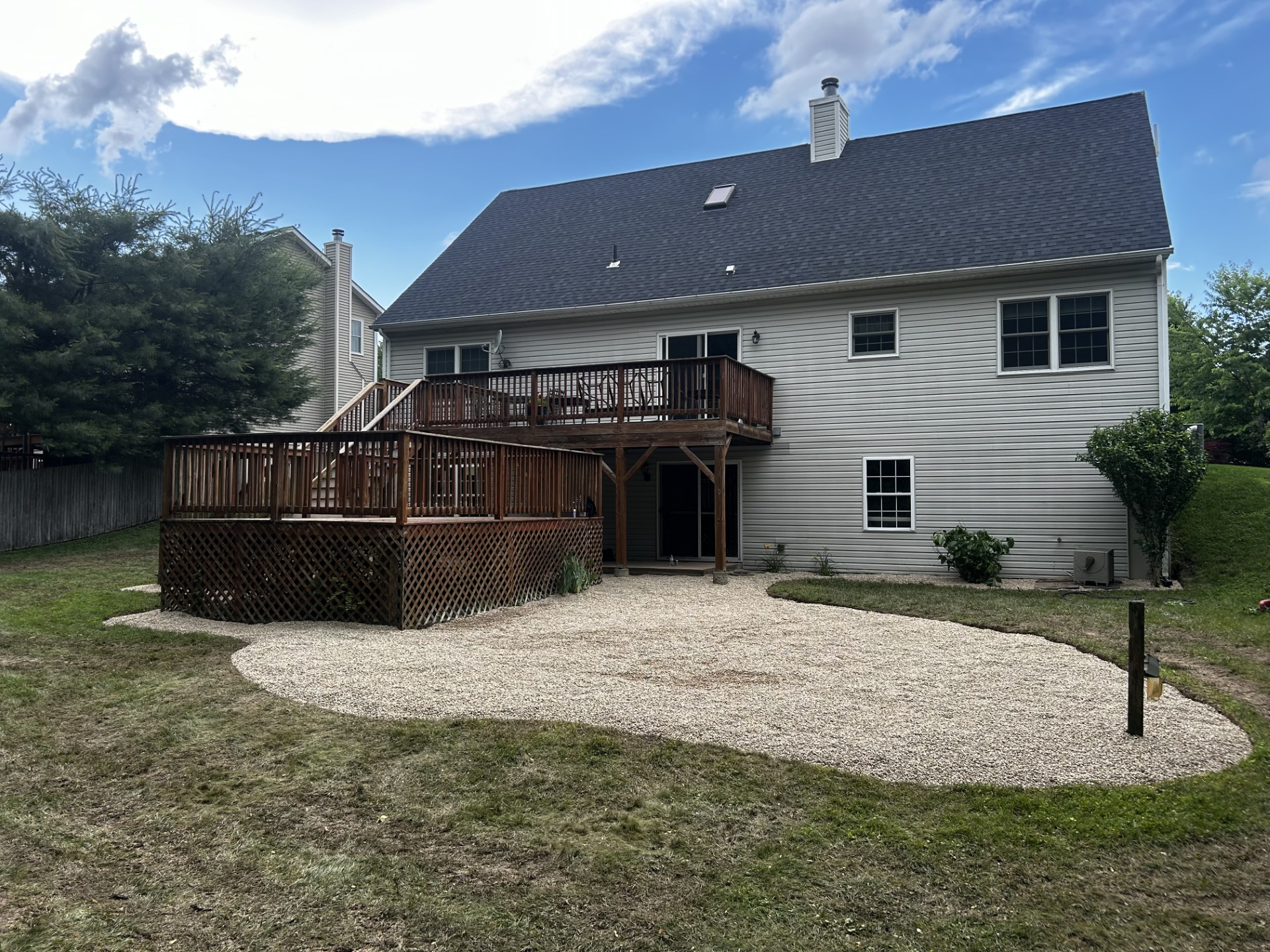 Back of a two-story house with a wooden deck and gravel patio in the backyard, blue sky.