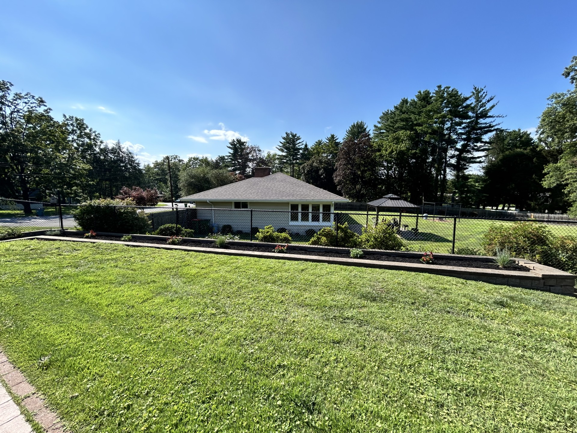 Small, white building with a dark roof surrounded by a garden bed and lawn under a blue sky.