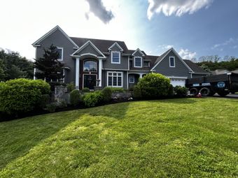 Large gray house with manicured lawn, landscaping, and a truck parked to the side on a sunny day.