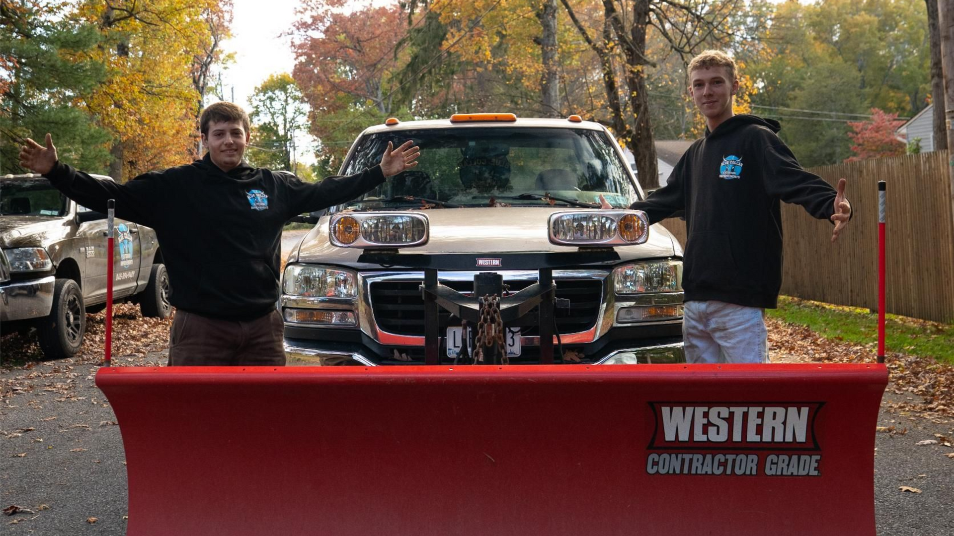 Two people stand with arms outstretched in front of a snow plow attached to a truck on a driveway.