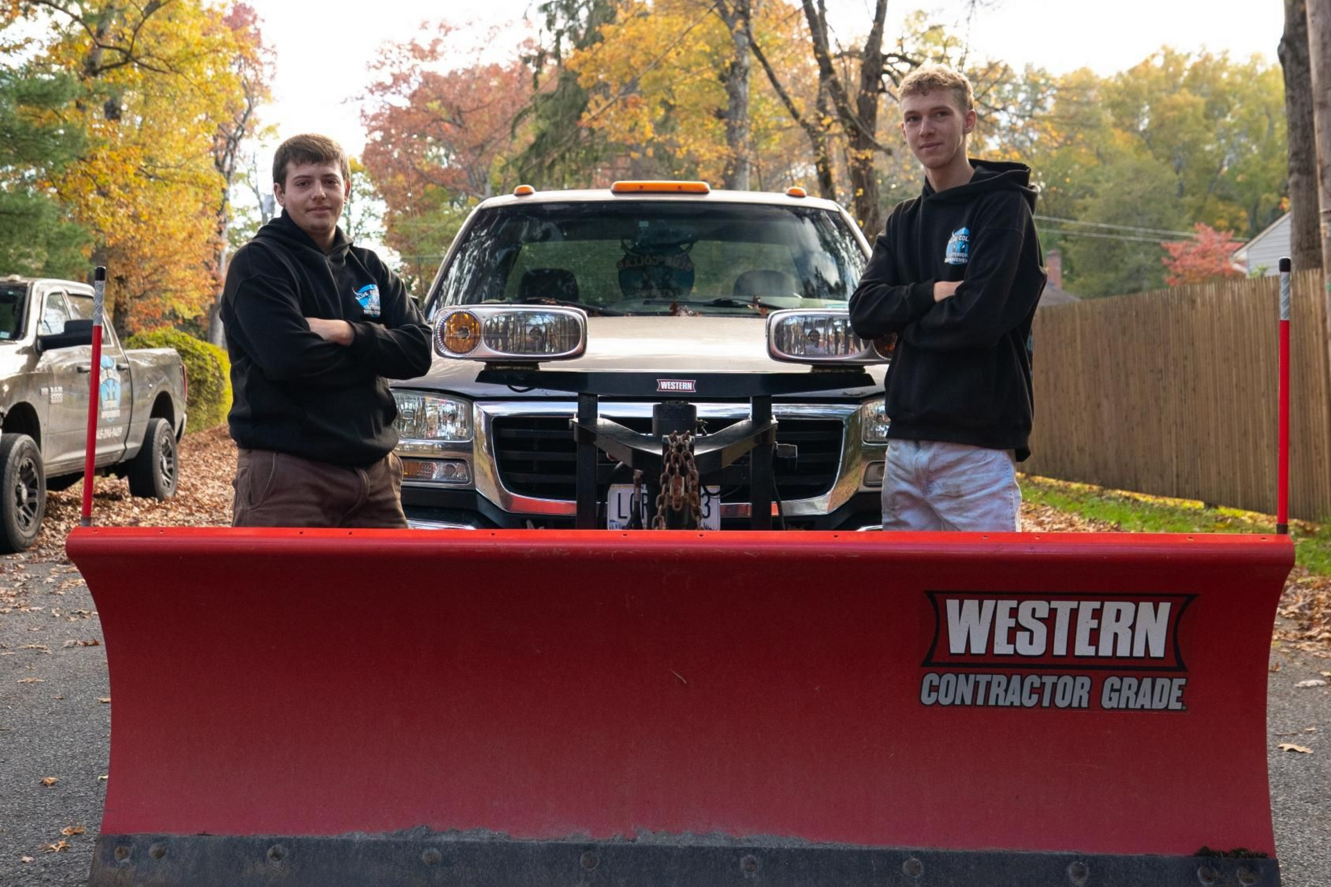 Two men stand in front of a snowplow truck with arms crossed. Autumn leaves in the background.