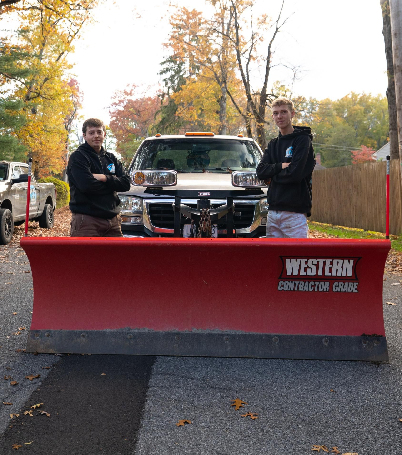 Two men stand in front of a snow plow attached to a truck. They are smiling and have their arms crossed. Autumn leaves are in the background.