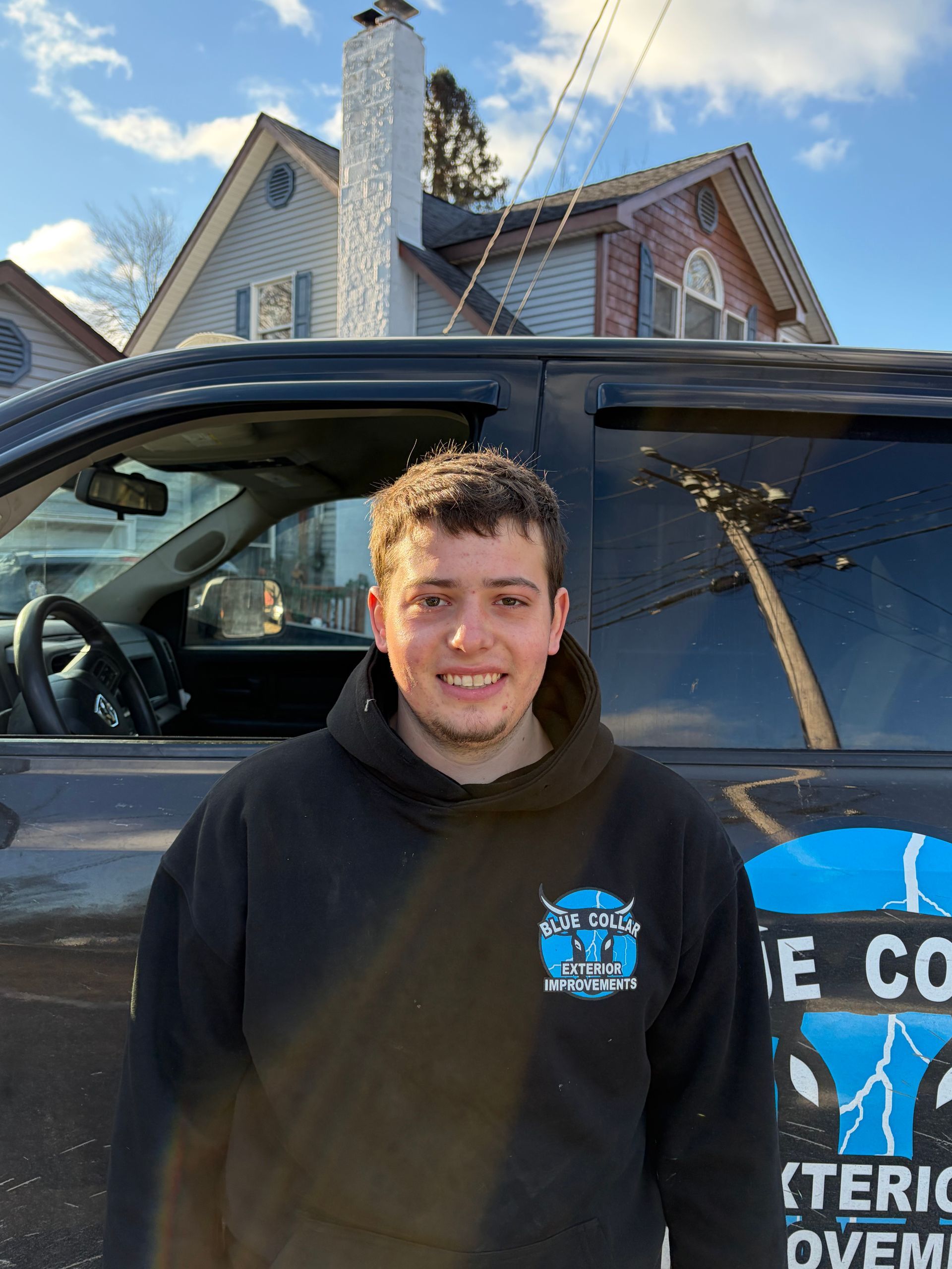 A person wearing a black hoodie stands in front of a truck, smiling, with a house in the background.
