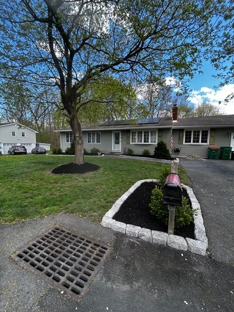 Suburban house with green lawn, tree, and driveway. Black mulch in beds, gray exterior, and mailbox.