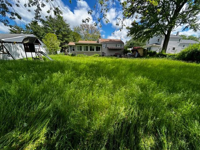 Grassy backyard with a house, sheds, and trees under a blue sky.