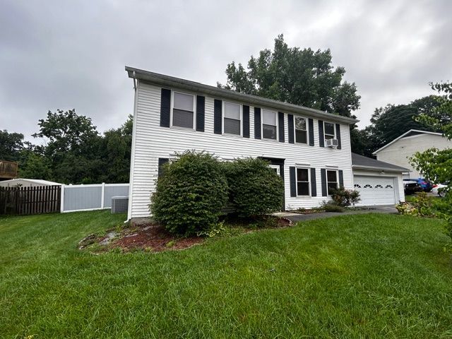 White two-story house with black shutters, green lawn, bushes, and a gray sky.