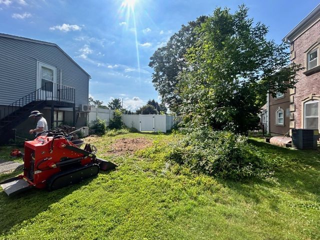 Man operates a red mini-skid steer clearing a grassy yard next to a two-story building on a sunny day.