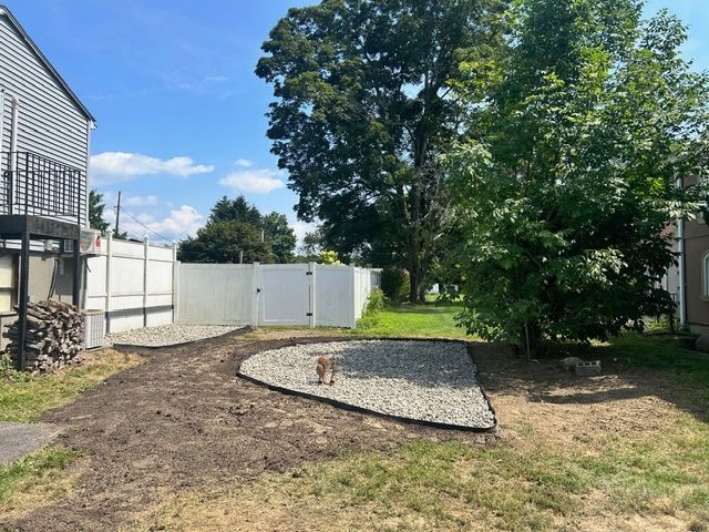 Backyard with gravel path, white fence, and small trees on a sunny day.