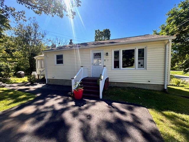 A single-story beige house with a red door, white railing, and a small porch. Bright sun shines.