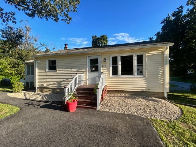 Yellow ranch-style house with dark asphalt driveway and gravel landscaping. Red flower pot on front steps. Sunny day.