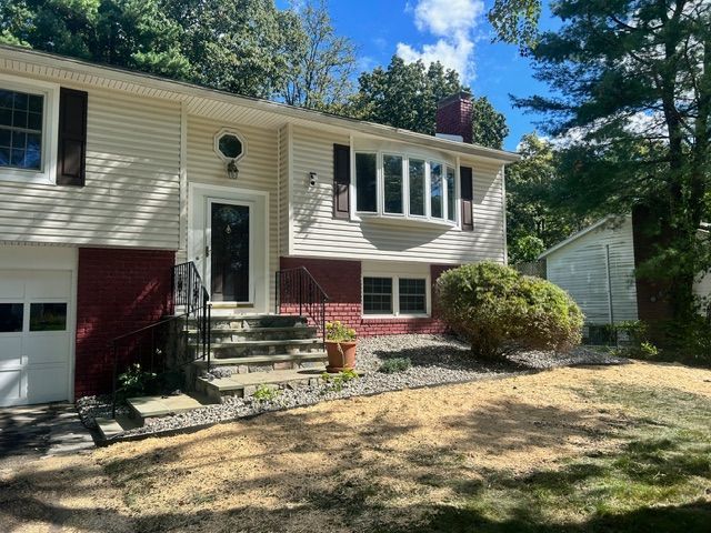 Tan and red split-level house with dark shutters, stone steps, and a bay window on a sunny day.