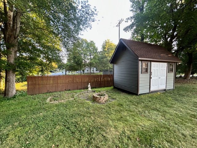 A backyard scene with a green shed, brown fence, and grassy area with a fire pit.
