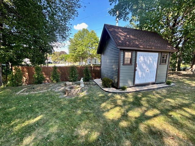 A small green shed with a brown roof sits in a grassy backyard, bordered by trees and a brown fence.