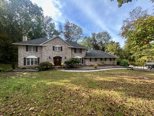 Two-story brick house with dark shutters, front lawn, and surrounding trees under a cloudy sky.