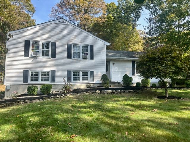White two-story house with black shutters, on a green lawn with a tree and bushes.