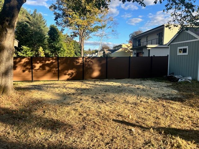 Backyard with a brown fence and a small shed. Empty, dirt ground with shadows. Bright sky.