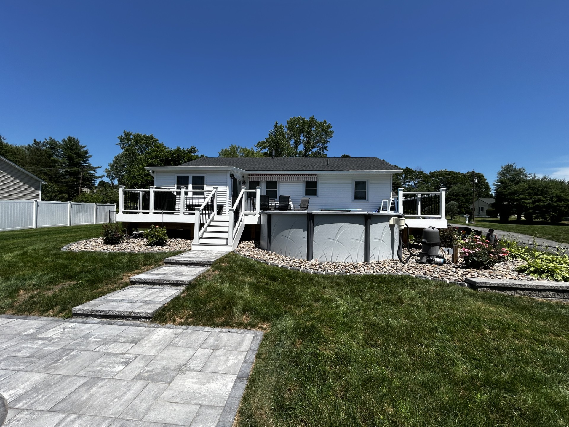 Backyard with white house, deck, above-ground pool, and stone patio under a blue sky.