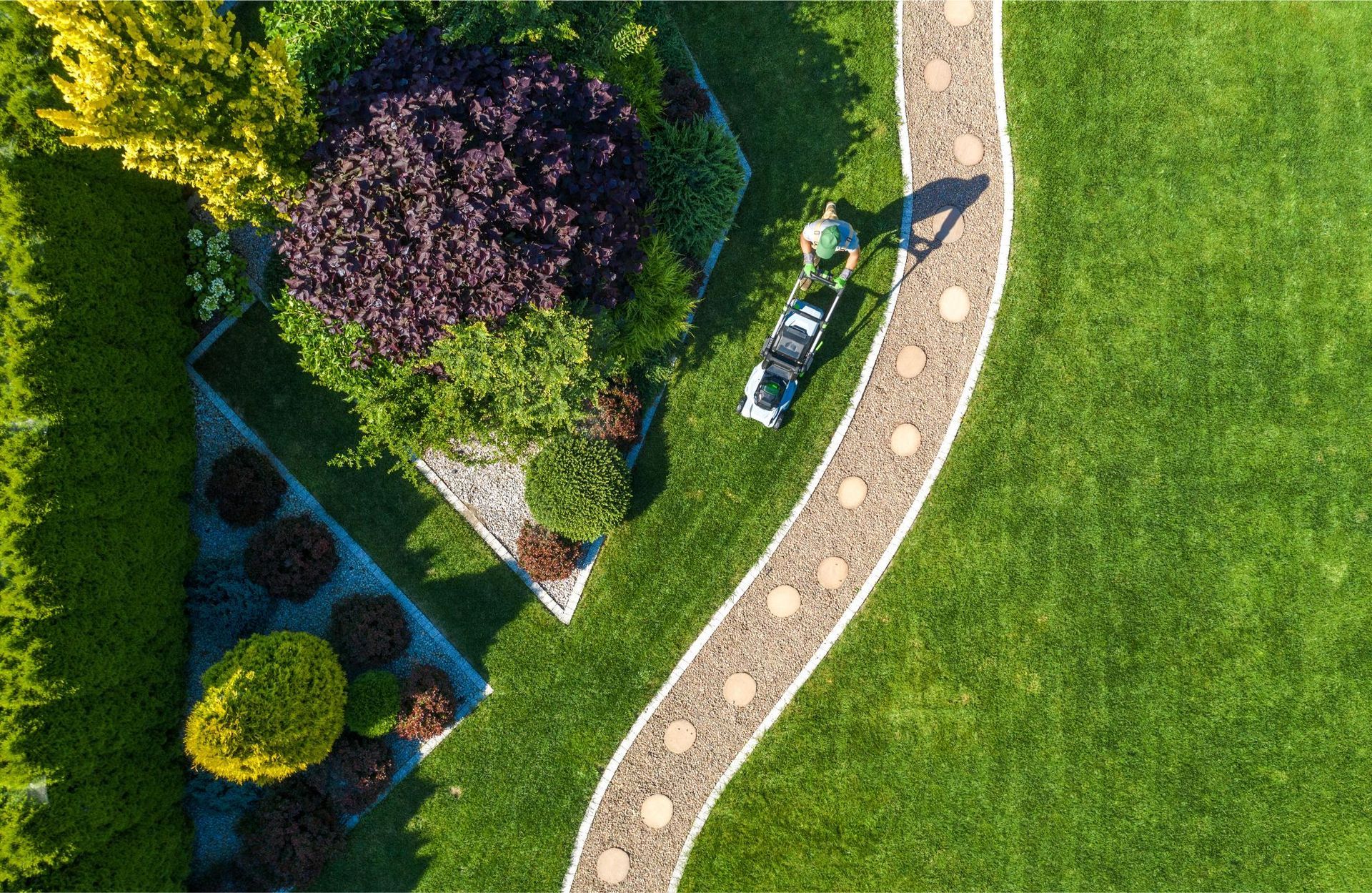 Overhead view of a person mowing green lawn with a winding stone path, bordered by shrubs and bushes.