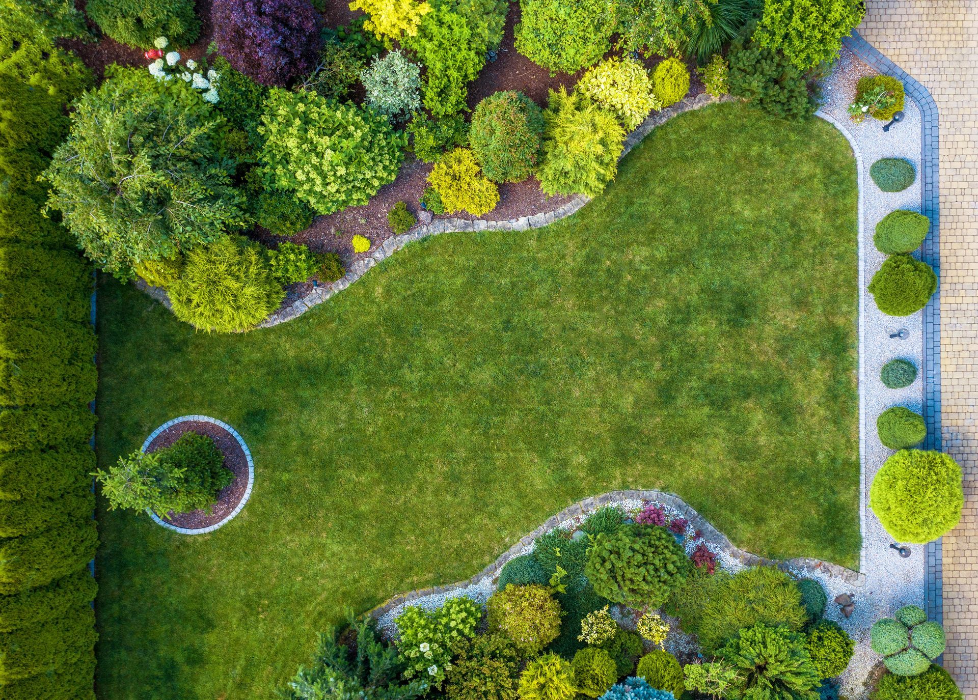 Aerial view of a green lawn bordered by colorful shrubs, a tree, and a stone pathway.