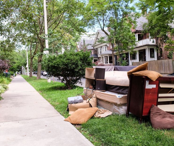 Pile of discarded furniture and debris on a grassy curb beside a sidewalk. Houses and trees line the street.